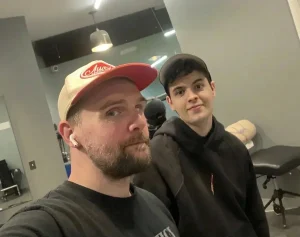 Two men sit in their apartment together taking a selfie and smiling.